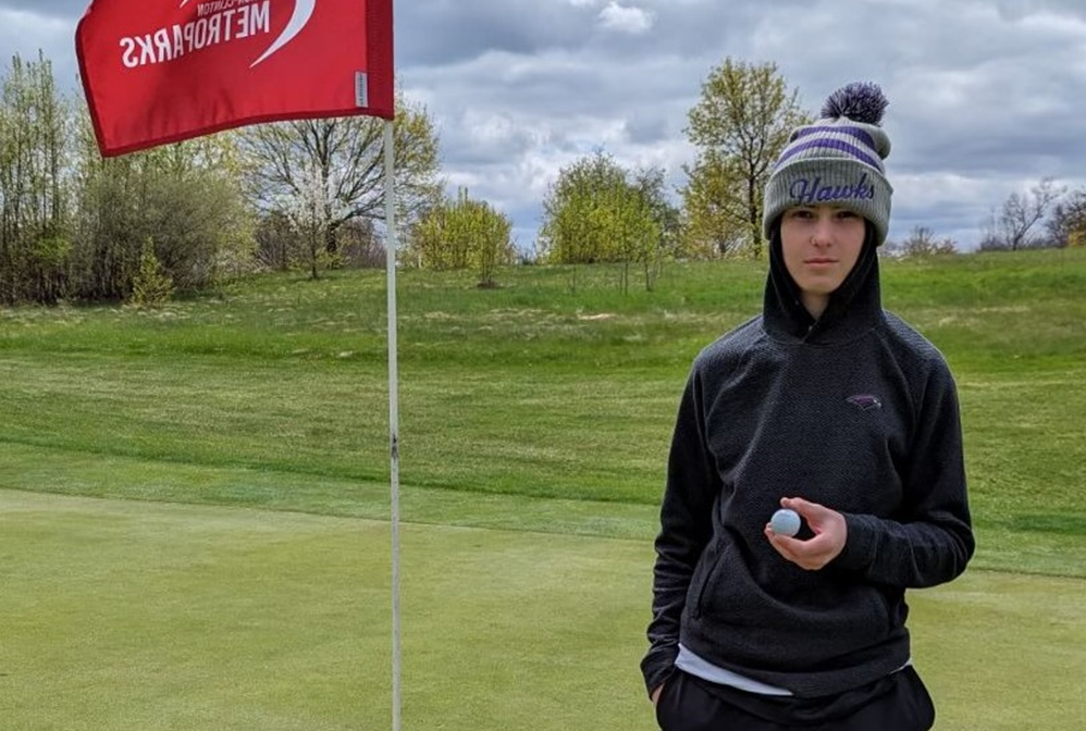 Bloomfield Hills’ Lucas Dostal shows the ball he drove into the hole next to him for a par-4 hole-in-one April 20 at Hudson Mills Metro Park. 