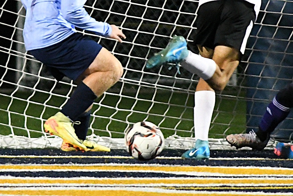 A soccer ball rests near the goal line amid multiple pairs of feet seeking to connect. 