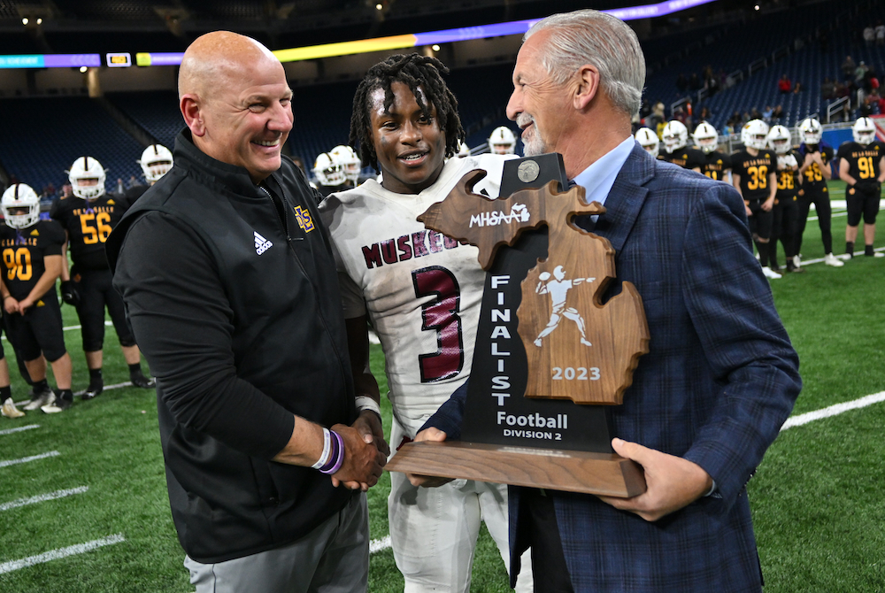 MHSAA Representative Council member Vic Michaels, far right, with the assistance of MHSAA Student Advisory Council member M'Khi Guy from Muskegon, presents Warren De La Salle Collegiate coach Dan Rohn the finalist trophy after the 2023 11-Player Football Final at Ford Field. 