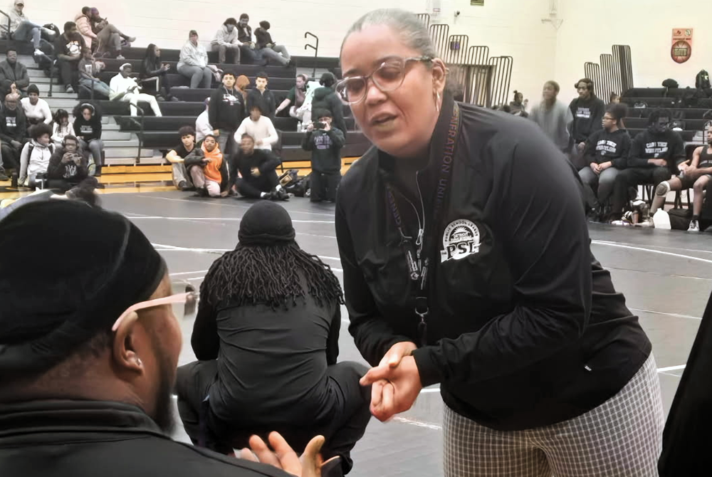 Anika McEvans communicates with someone sitting matside during a wrestling match.